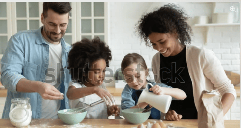 A mom, dad, and two young daughters in a modern home kitchen. They are cooking together, and the girl on the left is pouring a small pitcher into a bowl of flour.